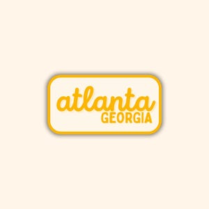 May include: A rectangular sticker with rounded corners, featuring the word "atlanta" in a yellow, cursive font above the word "GEORGIA" in a sans-serif font. The text is set against a cream-colored background, framed by a yellow border.