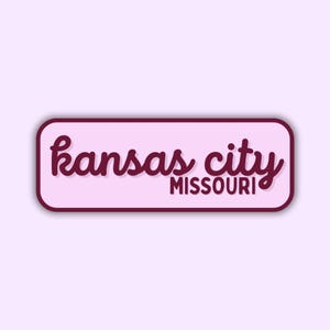 May include: A rectangular sticker with rounded corners, featuring the words "kansas city MISSOURI" in a cursive font. The text is in a dark red color, set against a light pink background, with a dark red border.