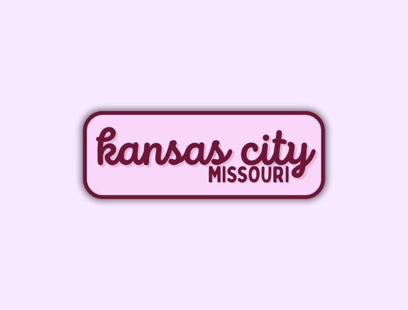 May include: A rectangular sticker with rounded corners, featuring the words "kansas city MISSOURI" in a cursive font. The text is in a dark red color, set against a light pink background, with a dark red border.