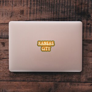 May include: A pink laptop computer with a sticker that says "KANSAS CITY" in a retro, colorful font.