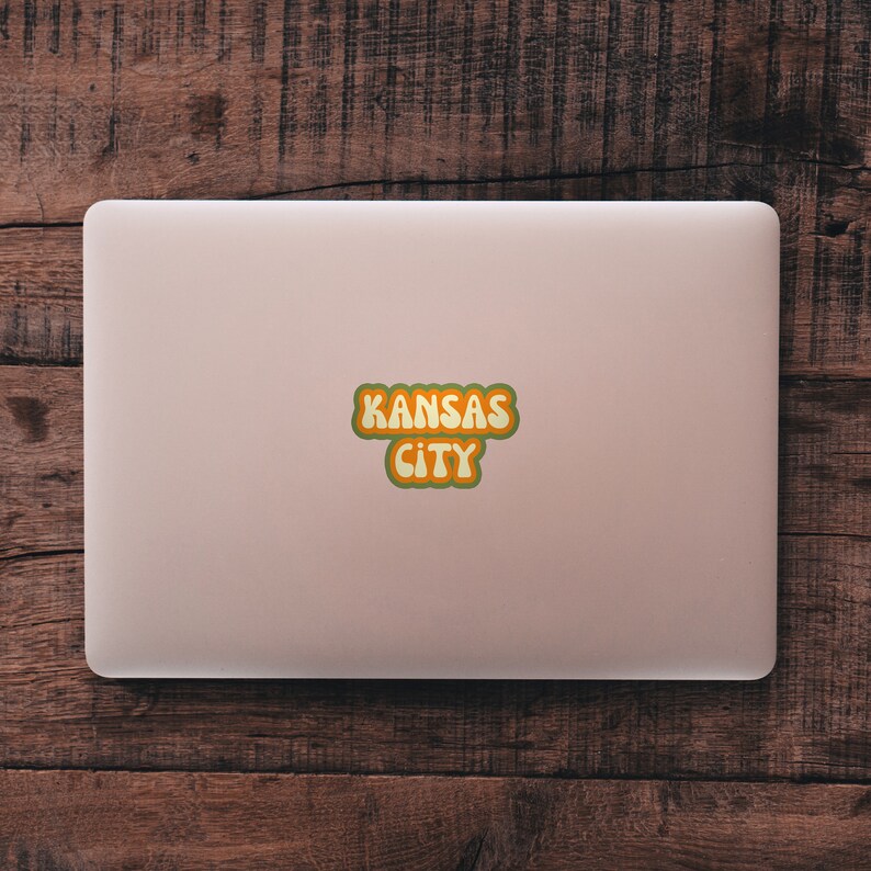 May include: A pink laptop computer with a sticker that says "KANSAS CITY" in a retro, colorful font.