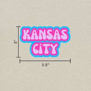 May include: A pink and blue sticker with the text "KANSAS CITY" in a retro font style. The sticker is 3.5 inches wide and 2 inches tall.