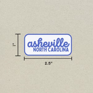 May include: A rectangular sticker with rounded corners, measuring 2.5 inches wide and 1 inch tall. The sticker features the word "asheville" in blue script above "NORTH CAROLINA" in blue block letters, all on a white background with a blue border.