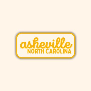 May include: A rectangular sticker with rounded corners, featuring the text "asheville NORTH CAROLINA" in a yellow, handwritten-style font. The text is set against a cream-colored background, with a yellow border, on a light beige background.