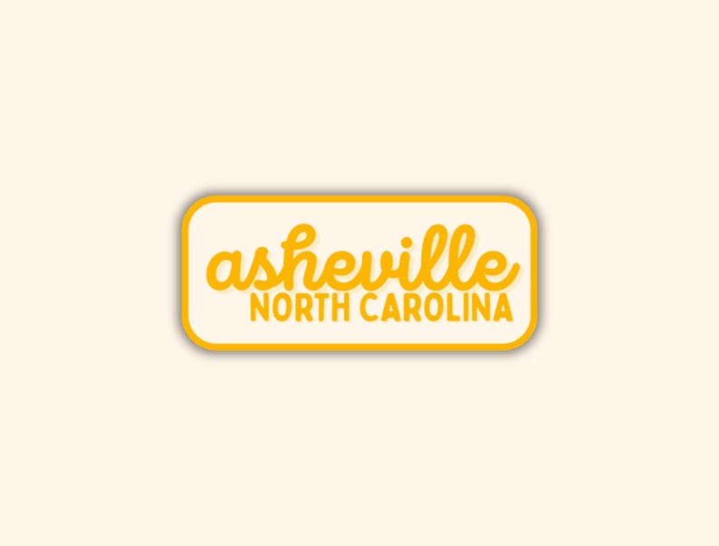 May include: A rectangular sticker with rounded corners, featuring the text "asheville NORTH CAROLINA" in a yellow, handwritten-style font. The text is set against a cream-colored background, with a yellow border, on a light beige background.