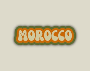 May include: A sticker with the word "MOROCCO" in a retro font. The text is cream-colored with an orange outline, set against a green border. The background is a light beige color.