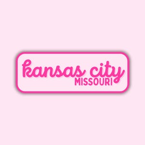 May include: A rectangular sticker with rounded corners, featuring the words "kansas city MISSOURI" in a vibrant pink script font. The text is set against a white background with a pink border, all on a light pink backdrop.