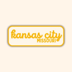 May include: A rectangular sticker with rounded corners. The sticker has a cream-colored background with a yellow border. The words "kansas city" are written in yellow cursive script above the word "MISSOURI" in a smaller, blocky yellow font.