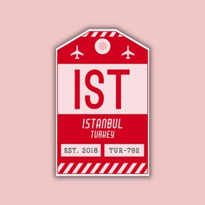 May include: A red and white luggage tag sticker with the letters "IST" in large, bold font. Below, the text reads "ISTANBUL TURKEY" and "EST. 2018". The tag features airplane icons and a striped design.