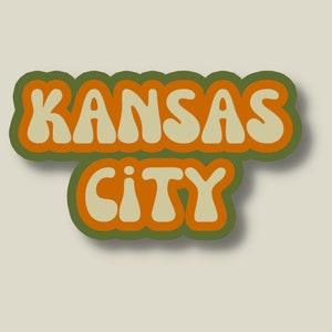 May include: A retro-style sticker with the text "KANSAS CITY" in orange and beige letters with a green outline.
