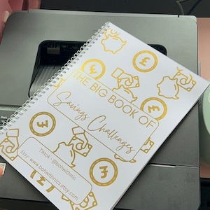May include: A white spiral bound notebook with the title "The Big Book of Savings Challenges" in gold lettering. The cover is decorated with gold coin and piggy bank illustrations.