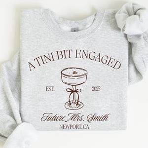 May include: A light gray sweatshirt with the text "A Tini Bit Engaged" and a cocktail illustration in brown. Below the illustration is "Future Mrs. Smith, Newport, CA" and the year "2025". The sweatshirt has a crew neck and long sleeves.