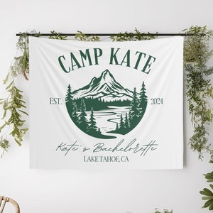 May include: White tapestry with green text that reads "Camp Kate Est. 2024" and a green illustration of a mountain range with a lake and trees. The text "Katie's Bachelorette Lake Tahoe, CA" is also included.