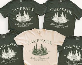 Peut inclure: Cinq t-shirts avec l'inscription "CAMP KATIE" et "Katie's Bachelorette LAKE TAHOE, CA" imprimée. Quatre chemises sont vert foncé et une est beige. Le motif représente une cabane et des arbres.