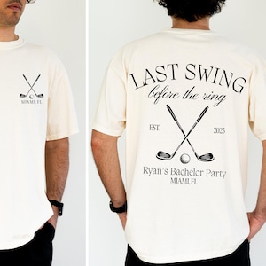 May include: Cream-colored t-shirt with a golf-themed design. The front features crossed golf clubs, a ball, and "Miami, FL." The back displays "Last Swing before the ring," crossed clubs, and "Ryan's Bachelor Party, Miami, FL."