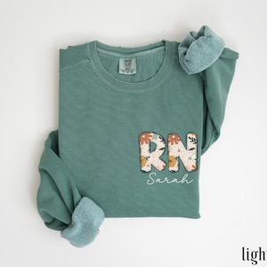 May include: Light green sweatshirt with a floral design that says "RN Sarah" on the front.