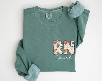 May include: Light green sweatshirt with a floral design that says "RN Sarah" on the front.