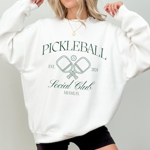 May include: White sweatshirt with a green graphic design that says "Pickleball Social Club" with a pickleball paddle and ball design. The text also says "Est. 2024" and "Miami, FL".