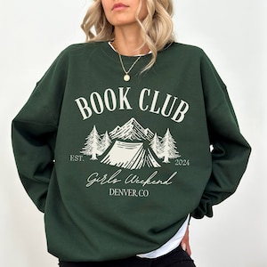 May include: Green sweatshirt with a white graphic design that says "Book Club" with a mountain scene and a tent. The text below the graphic says "Girls Weekend Denver, CO" and "Est. 2024".