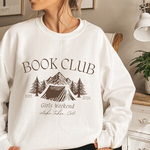 a woman wearing a book club sweatshirt standing in front of a bed