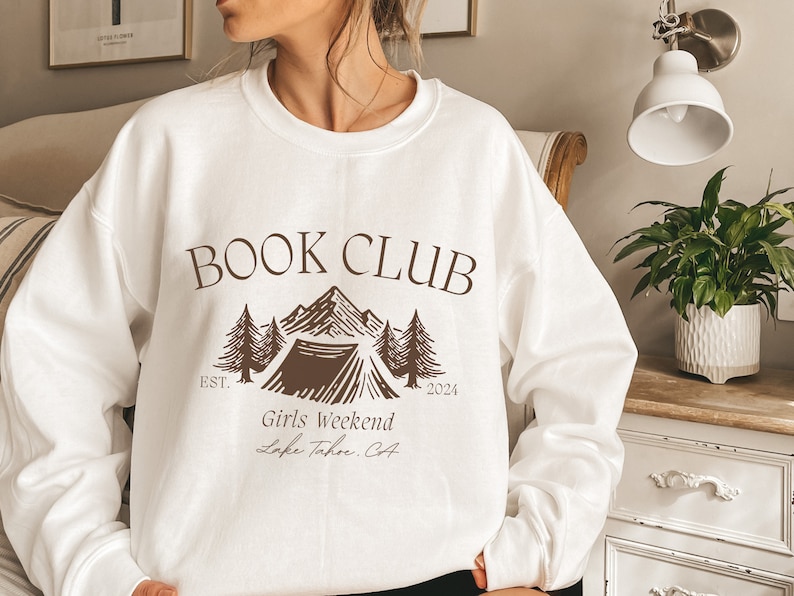 a woman wearing a book club sweatshirt standing in front of a bed