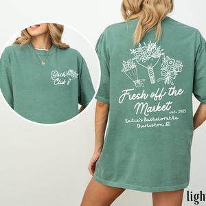 Może przedstawiać: Jasnozielony T-shirt z białym nadrukiem bukiet&oacute;w kwiat&oacute;w i tekstem "Fresh off the Market est. 2025 Katie's Bachelorette Charleston, SC"