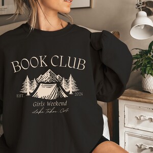 a woman holding a book club sweatshirt while sitting on a bed