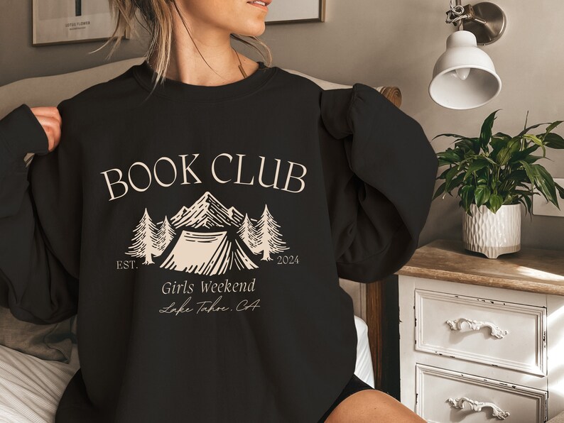 a woman holding a book club sweatshirt while sitting on a bed