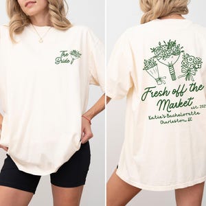 Może przedstawiać: Biały t-shirt z zielonym nadrukiem bukietu kwiat&oacute;w i tekstem "Fresh off the Market est. 2025 Katie's Bachelorette Charleston SC". Prz&oacute;d koszulki zawiera napis "The Bride" z małym nadrukiem bukietu kwiat&oacute;w.
