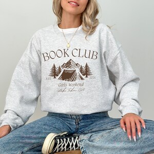 a woman sitting on the floor wearing a book club sweatshirt