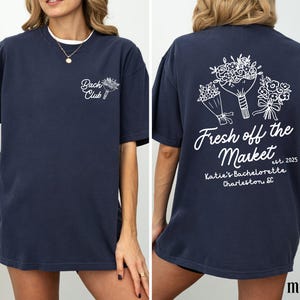 Może przedstawiać: Koszulka w kolorze granatowym z białym nadrukiem i kwiatowymi grafikami. Z przodu koszulka ma napis "Bach Club", a z tyłu "Fresh off the Market est. 2025 Katie's Bachelorette Charleston, SC".