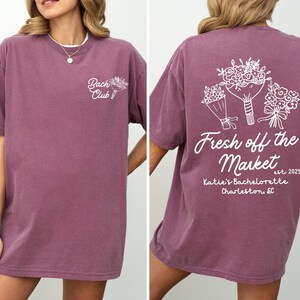 Może przedstawiać: Fioletowy T-shirt z napisem "Bach Club" po lewej stronie i wzorem kwiatowym z napisem "Fresh off the Market est. 2025 Katie's Bachelorette Charleston, SC" po prawej stronie.