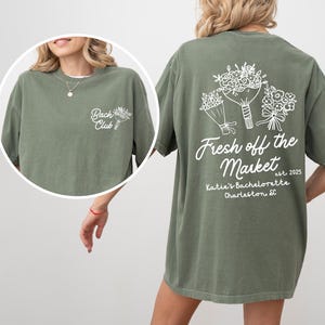 Może przedstawiać: Zielona koszulka z białym napisem "Fresh off the Market est. 2025 Katie's Bachelorette Charleston SC" i grafiką trzech bukiet&oacute;w kwiat&oacute;w. Prz&oacute;d koszulki ma biały napis "Bach Club" z małą grafiką kwiatu.