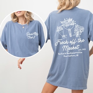 Może przedstawiać: Jasnoniebieska koszulka z białym nadrukiem kwiat&oacute;w i tekstem "Fresh off the Market est. 2025 Katie's Bachelorette Charleston, SC".