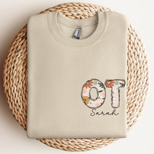 May include: A beige sweatshirt with the letters "OT" in floral print and the name "Sarah" written below.