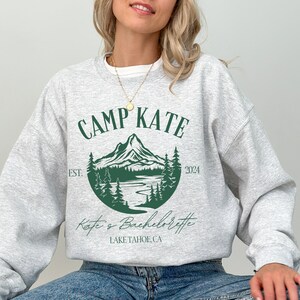 May include: Gray sweatshirt with a green graphic design that says "Camp Kate" with a mountain, trees, and a river. The graphic also says "Est. 2024" and "Kate's Bachelorette Lake Tahoe, CA".