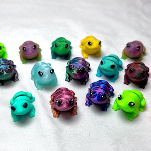 Puede incluir: Una colección de 18 pequeñas figuritas de rana en varios colores, incluyendo verde, azul, morado, rosa, naranja y amarillo. Las ranas están hechas de un material liso y brillante y tienen ojos negros.