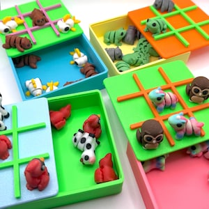 May include: Five colourful tic-tac-toe game boxes with different animal themed pieces. The boxes are green, orange, pink, yellow, and blue. The pieces are made of plastic and are in various colours.