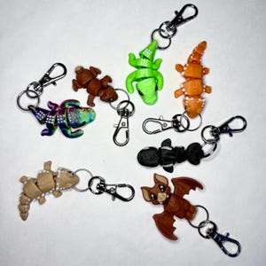 May include: A set of eight colorful animal keychains. The keychains feature a variety of animals, including a chameleon, a bat, a crocodile, and a lizard. The keychains are made of a soft, pliable material and are perfect for adding a touch of whimsy to your keys or bag.