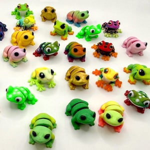 Puede incluir: Una colección de figuras de ranas coloridas en varios tamaños y diseños. Las ranas son en tonos de verde, amarillo, rosa y dorado, algunas con patrones y detalles. Las figuras están dispuestas sobre una superficie blanca.