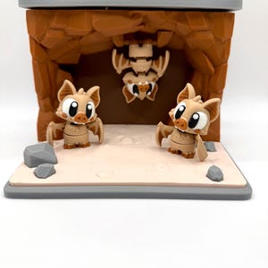 May include: A diorama featuring three cartoon bats in a cave setting. Two bats stand on a sandy base, while a third hangs upside down from the cave ceiling. The cave is brown, and the base is gray.