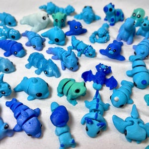 May include: A collection of small, colorful, blue, green, and white figurines. The figurines are made of plastic and have a variety of animal and creature designs, including a frog, a bat, a dolphin, a dragon, and a caterpillar.