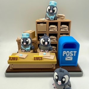 Puede incluir: Un diorama caprichoso impreso en 3D con palomas cartero. La escena incluye un buzón azul, un escritorio con sobres y estantes con paquetes. Las palomas llevan sombreros azules y bolsas de mensajero. La paleta de colores es principalmente gris, azul y beige.