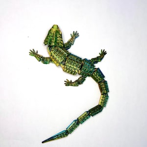 May include: A green and blue articulated lizard figurine with a segmented tail. The lizard is posed in a walking position with its head tilted up.