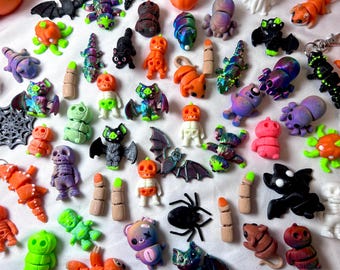 Peut inclure: Une collection de petites figurines articulées sur le thème d'Halloween, incluant des citrouilles, squelettes, chauves-souris, araignées et doigts dans des couleurs variées comme l'orange, le vert, le violet et le noir. Ces miniatures ont des articulations mobiles.