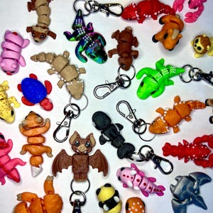 May include: A collection of colorful, small, animal-shaped keychains. The keychains are made of a variety of materials, including plastic, metal, and fabric. They feature a variety of animals, including a sloth, a bat, a lizard, a crab, a bee, a lobster, a fish, and a dragon.