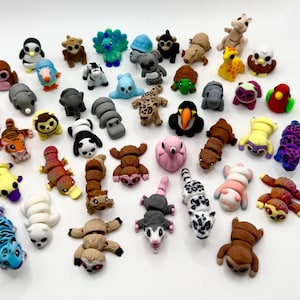 Pode incluir: Uma coleção de figuras de animais coloridas, incluindo um pinguim, um macaco, um coala e uma girafa. As figuras são feitas de um material macio e estão em várias cores, como azul, castanho e amarelo. A coleção inclui uma variedade de animais.