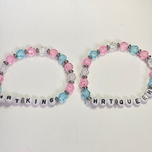 May include: Two beaded bracelets with alternating pink, blue, and white beads. The bracelets spell out "HRT KING" and "HRT QUEEN" in black letters on white beads.