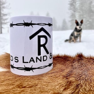 Puede incluir: Taza de cer&aacute;mica blanca con un dise&ntilde;o de alambre de p&uacute;as negro y el texto "R DS LAND &" impreso en ella. La taza est&aacute; sobre una superficie de piel marr&oacute;n.