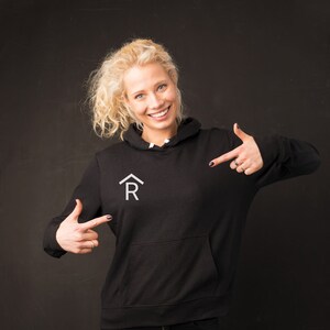 Puede incluir: Una mujer con una sudadera con capucha negra con un logotipo blanco de una casa y la letra R. Est&aacute; se&ntilde;alando el logotipo con ambas manos.